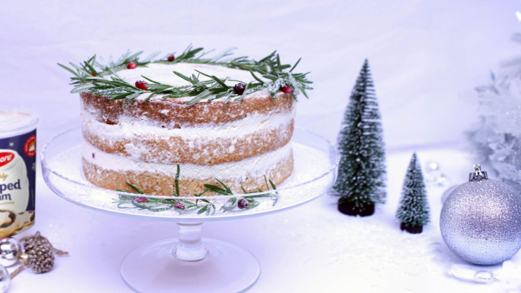 christmas layered cake topped with rosemary on a glass cake stand beside a carton of avonmore freshly whipped cream