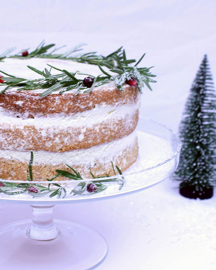 christmas layered cake topped with rosemary on a glass cake stand beside a carton of avonmore freshly whipped cream
